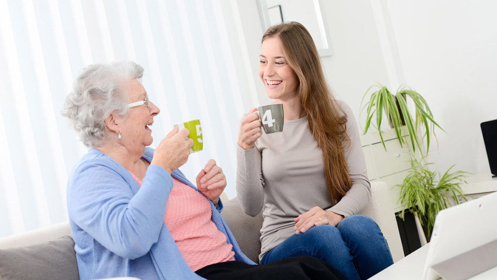 weibliche Pflegekraft und ältere Dame sitzen lachend auf der Couch mit zwei Tassen in der Hand.