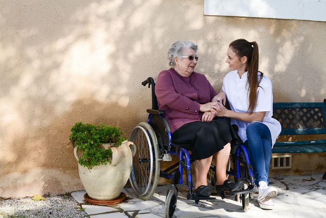 ältere Frau im Rollstuhl und Pflegekraft, die neben ihr auf einer alten blauen Parkbank sitzt. Beide sind zueinander gebeugt, lachen sich gegenseitig an und halten sich an den Händen.