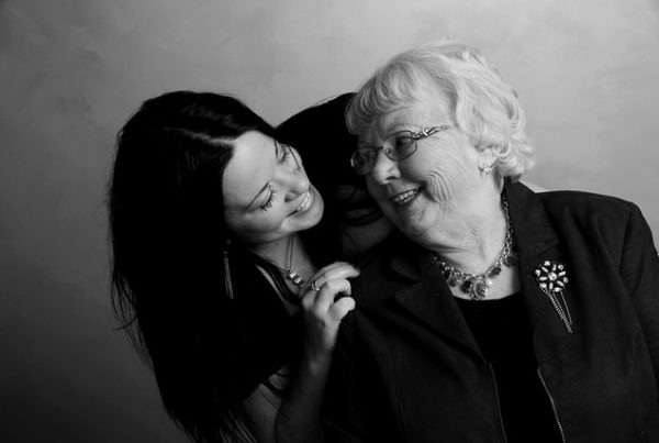 Großmutter an Enkelin angelehnt, die sich über ihre rechte Schulter beugt. Beide lachen sich an.