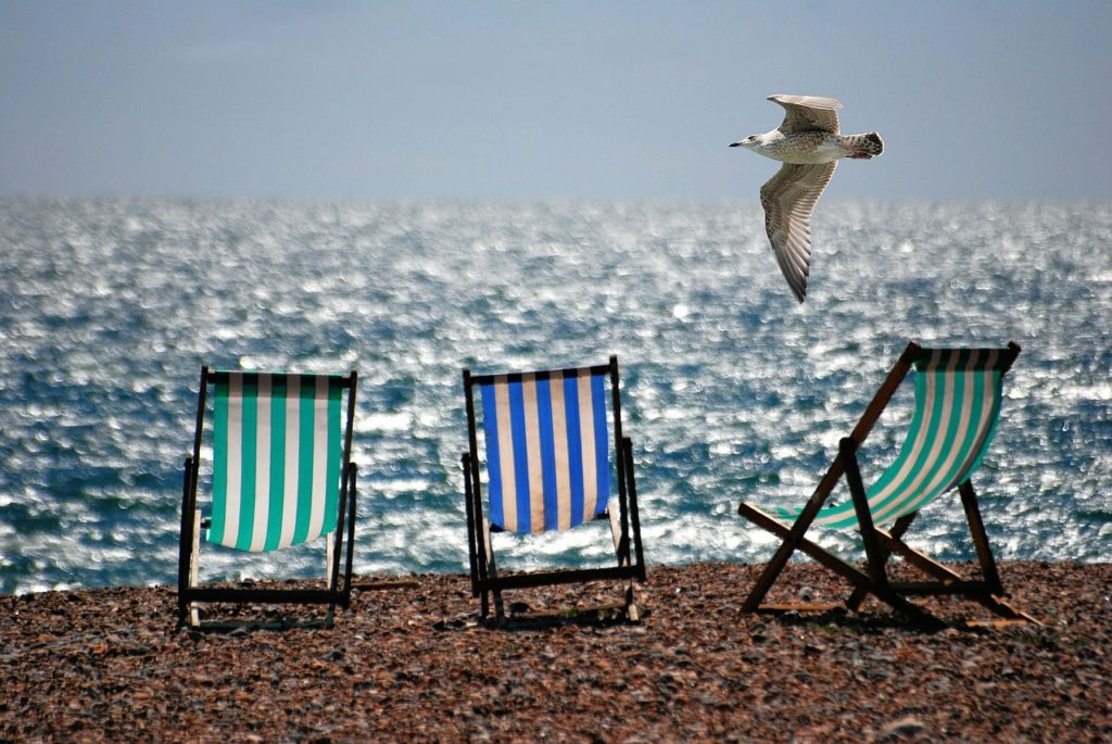 3 gestreifte Liegestühle, die in einer Reihe am Strand vorm Meer stehen. Möwe fliegt oben drüber.