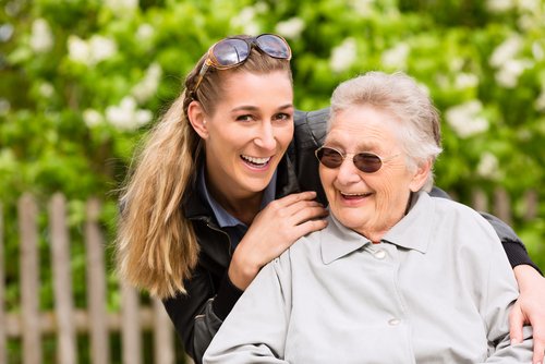 sitzende ältere Frau, dahinter weibliche Pflegekraft, die sich über die rechte Schulter beugt. Beide lachen und sind im Garten.