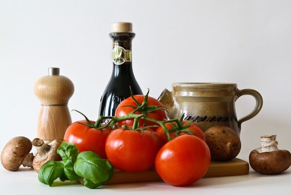 Tomate, Pilze und Basilikum auf einem Brett. Dahinter stehen Essig, Öl und eine Pfeffermühle.