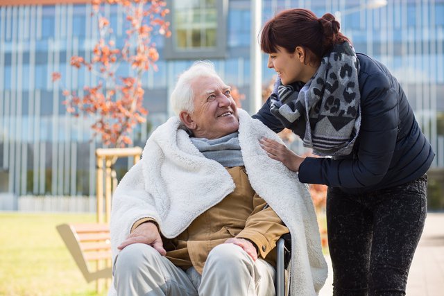 älterer Herr im Rollstuhl, der zugedeckt ist. Daneben Pflegekraft, die die Deckte zurechtrückt. Beide lachen sich gegenseitig an und sind im Freien.