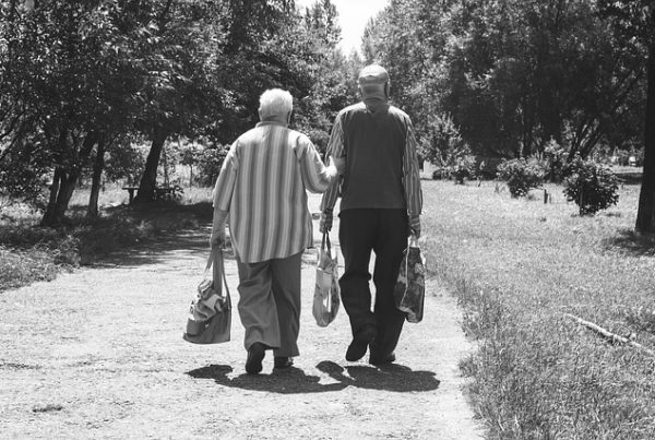 schwarz-weiß Foto von zwei älteren Herren, die auf einem Feldweg gehen. Der Rechte hat zwei Einkaufstüten in der Hand, der Linke hat in einer Hand eine Einkaufstüte und mit der anderen Hand hält er sich bei seinem Begleiter an.